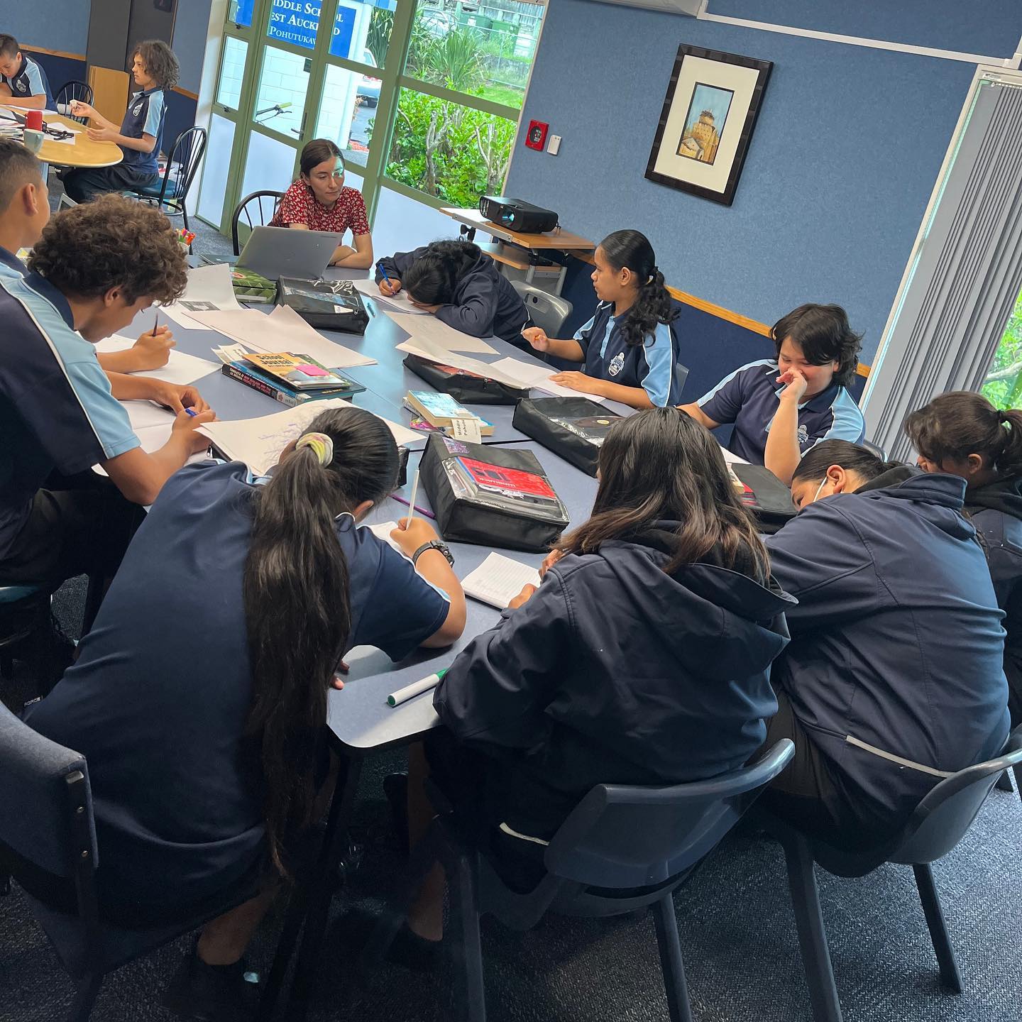 A small group of students sit around a shared table in a classroom.
			They are writing in their notebooks.