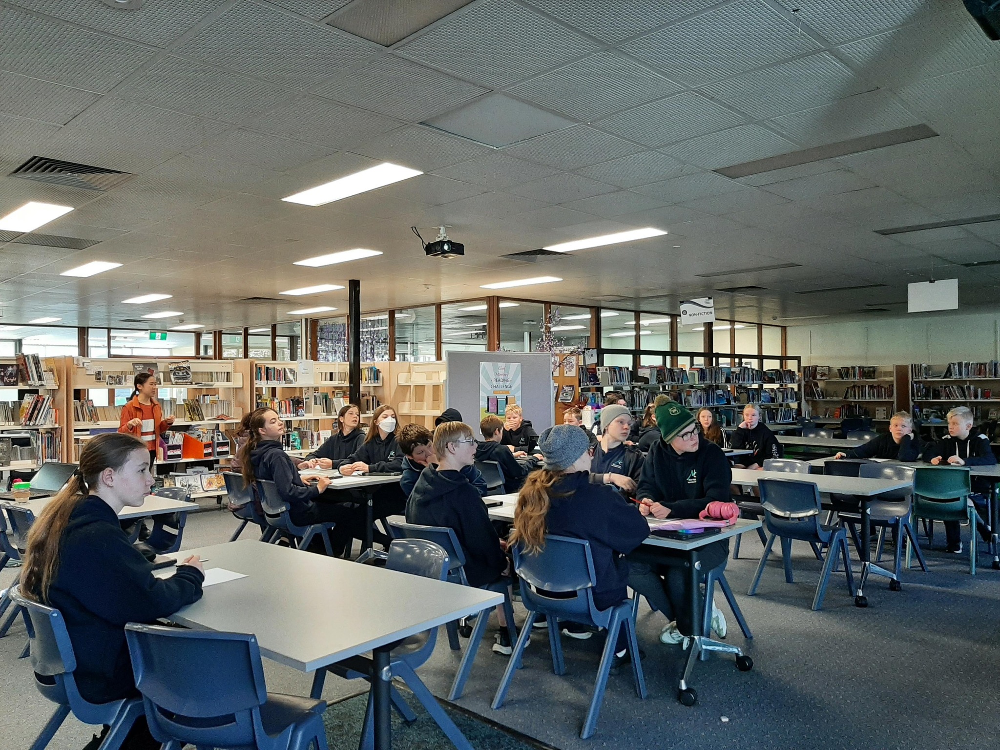 Lots of students sit at desks in a classroom, looking at the teacher
			to the right (out of shot). There are lots of shelves with books in the
			background.