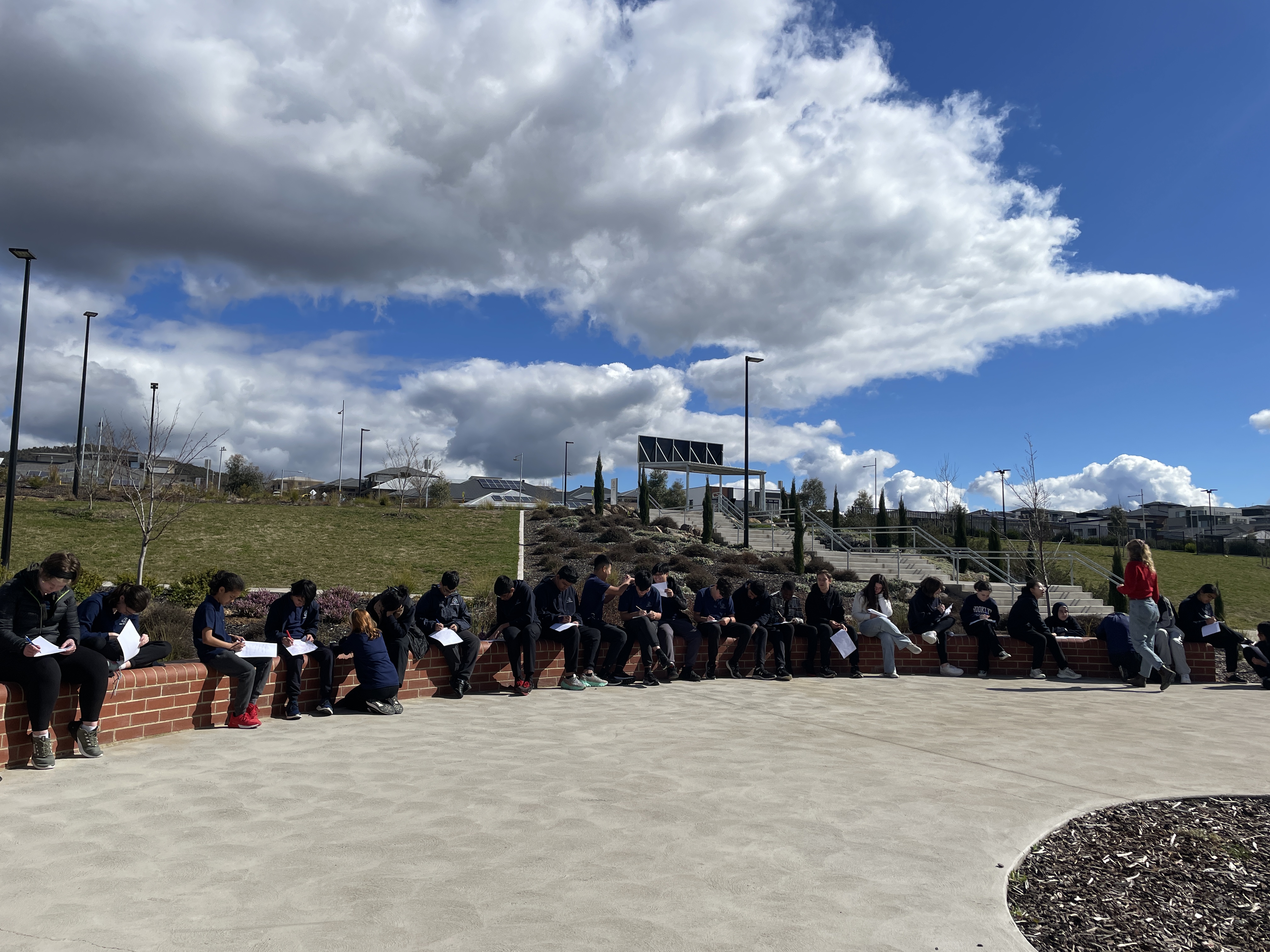 Students are sitting outside on a long, low brick wall. The sky is a
			vivid blue, with clouds. The students are busily writing in their
			notebooks.